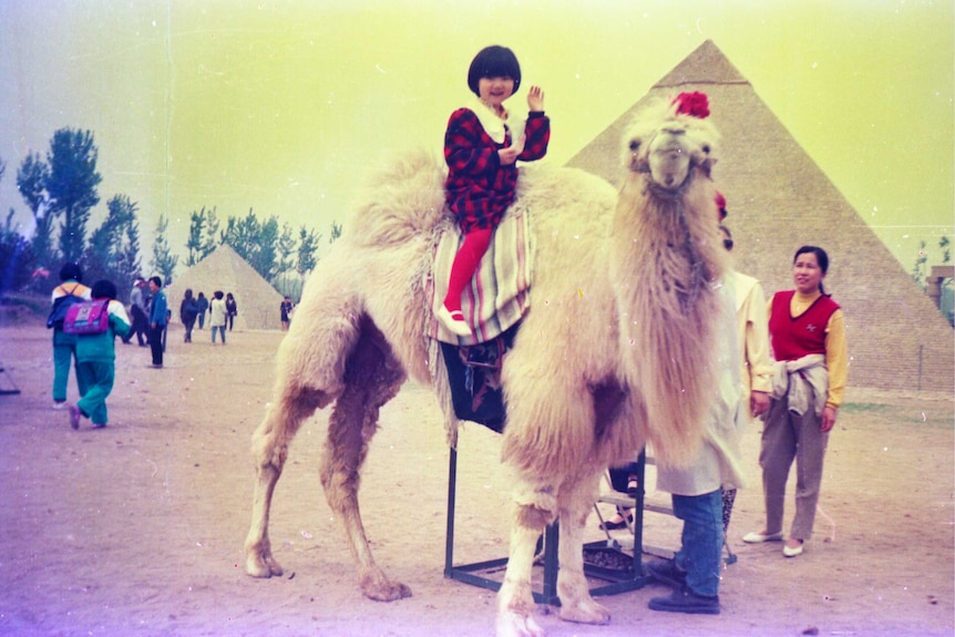 You view a young girl waving to the camera as she sits atop a white camel in front of a re-creation of an Egyptian pyramid.