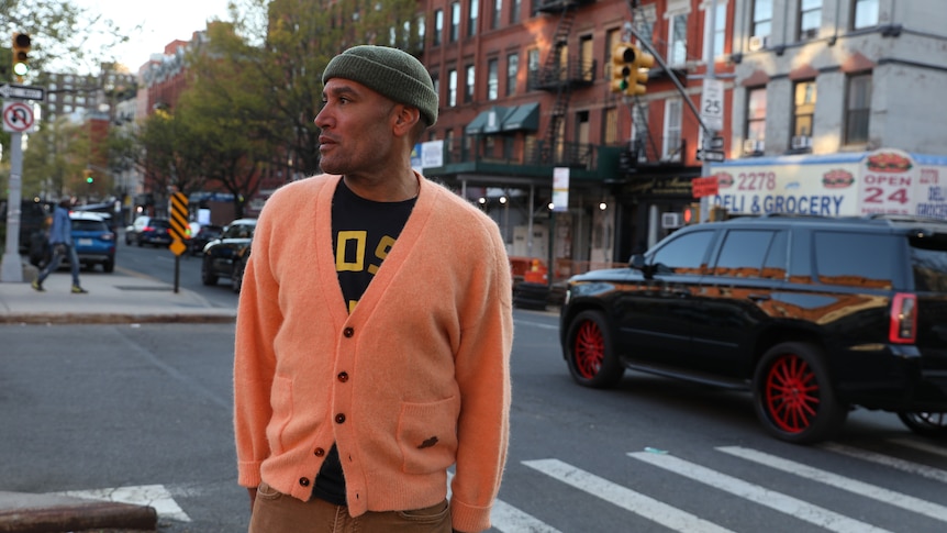 Ben Harper wears a wool cap and cardigan. He stands on a city street looking away from the camera.