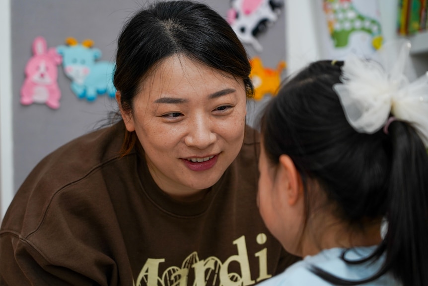 An Asian woman wearing a brown jumper smiles at a little girl wearing a white ribbon in her hair.