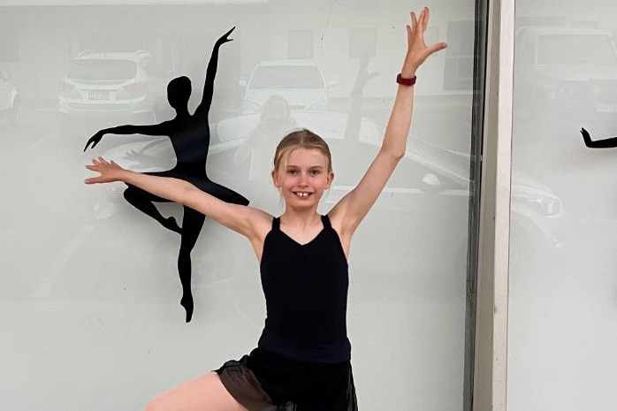 A young blonde ballerina in a black leotard and black skirt performs a ballet move in front of her dance school in Longreach.