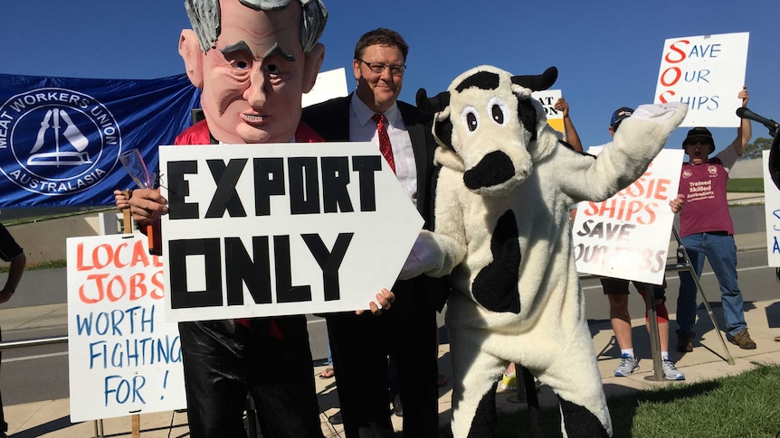 Man stands with person in cow suit and man with papier mache head at protest