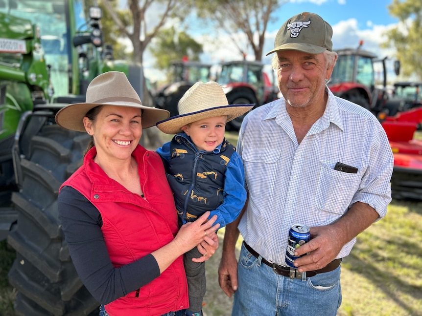 a woman and young son and a granddad smile in front of a tractor