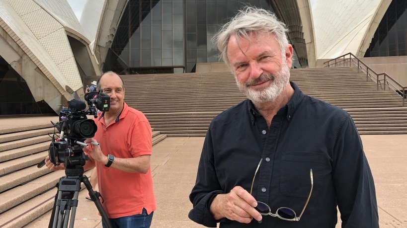 Cameraman holding camera on tripod standing behind Neill with Opera House in background.
