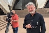 Cameraman holding camera on tripod standing behind Neill with Opera House in background.