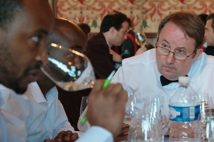 Tinashe Nyamudoka is seen at left holding a glass of red wine to his nose while a man in a white jacket watches him intently.