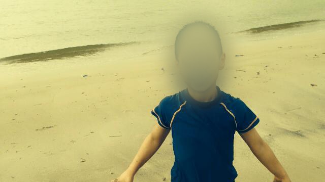 A boy stands on a beach. His face is blurred.
