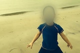 A boy stands on a beach. His face is blurred.