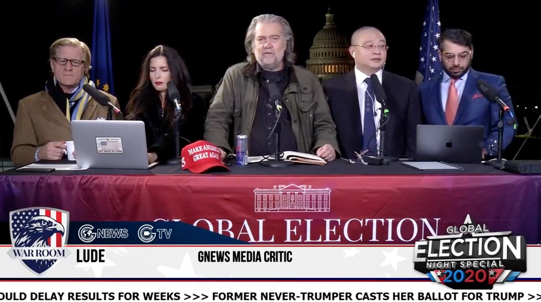 Five people sit behind a panel table presenting a broadcast with the US Capitol Building in the background