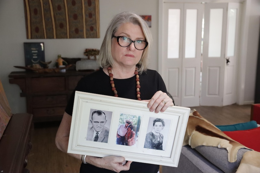 A sad-looking woman seated on a couch holding a picture frame featuring three photos of her late parents.
