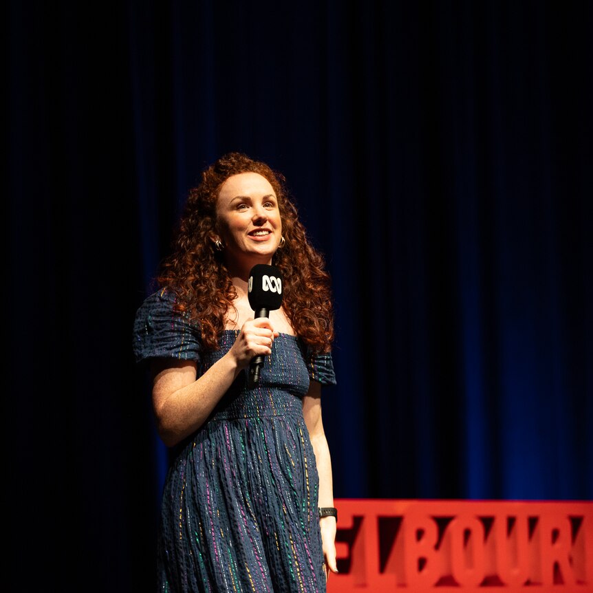 Catherine Bohart  on stage with a red Comedy Festival sign and dark blue stage curtains in the background.