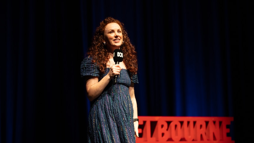 Catherine Bohart  on stage with a red Comedy Festival sign and dark blue stage curtains in the background.