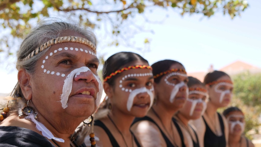 Yamatji women, getting ready to dance with their faces traditionally painted sit in a line under a tree.