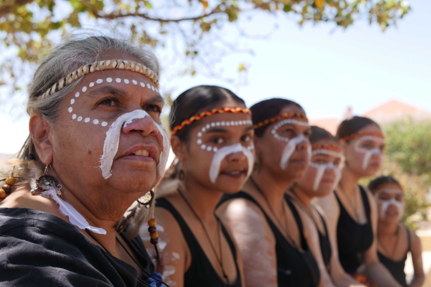 Yamatji women, getting ready to dance with their faces traditionally painted sit in a line under a tree.