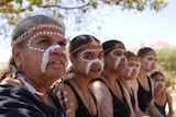Yamatji women, getting ready to dance with their faces traditionally painted sit in a line under a tree.