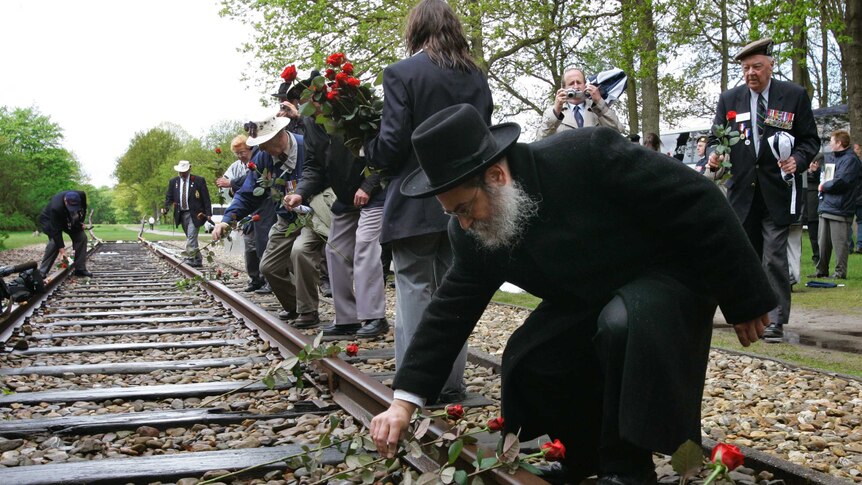 A rabbi is surrounded by WWII veterans placing roses on a section of railroad track outside of a former concentration camp.