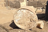 A large teracotta jar filled with a white substance rests on its side on some large bricks/stones in the desert amid rubble