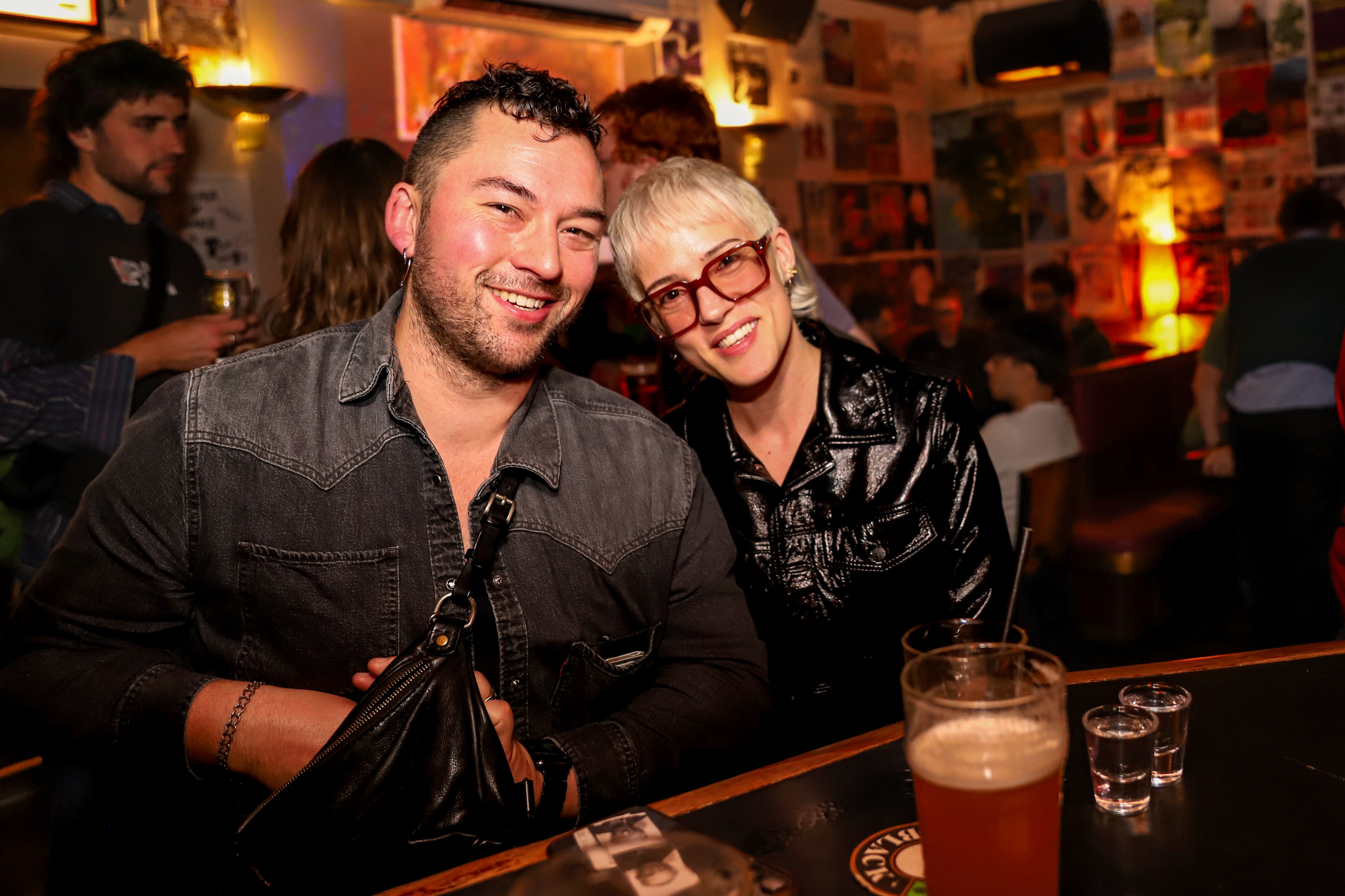 Um homem e uma mulher sorriem enquanto se sentam no bar de um pub movimentado e mal iluminado. 