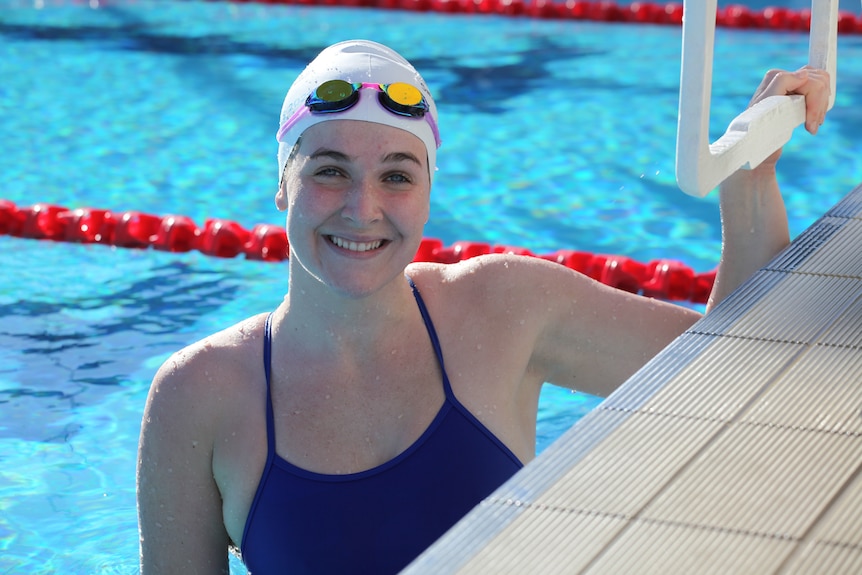 A woman holds onto the blocks at the swimming pool from the water and smiles at the camera.