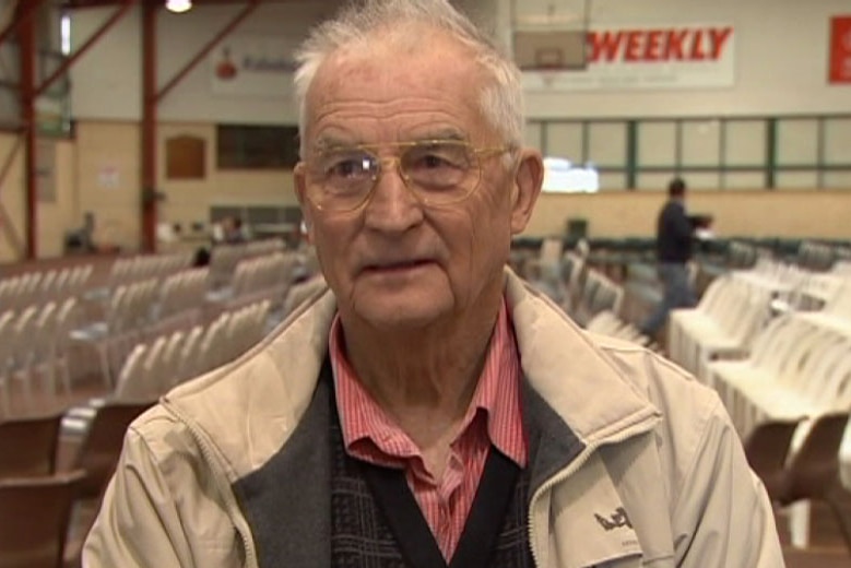 Colin stands in a large sports centre filled with chairs.