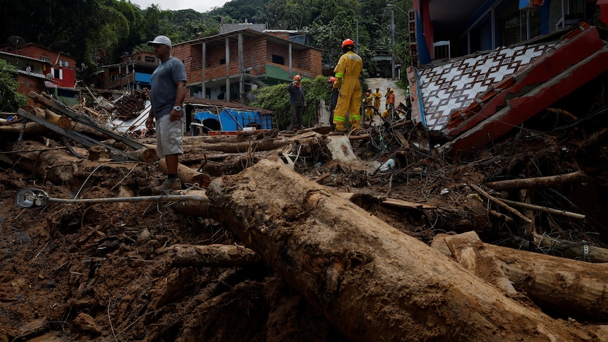 Debris across the ground with buildings in the background as people stand.