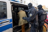 Two masked police in riot gear push a man in a tweed jacket into the back of a police van. 