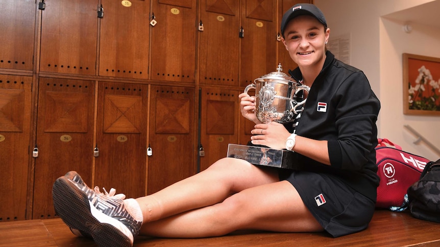 A tennis player sits on a bench in the locker room with the trophy after winning the French Open.