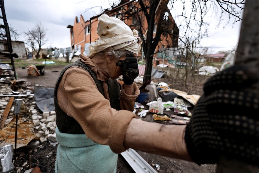 Elderly woman cries as she uses arm to lean against pole outside destroyed home.