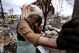 Elderly woman cries as she uses arm to lean against pole outside destroyed home.