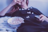 A woman with red hair lies on a bed holding a remote with popcorn spilling from a bowl