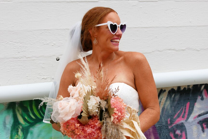 A woman in a wedding dress and heart-shaped sunglasses holds a large bouquet of flowers