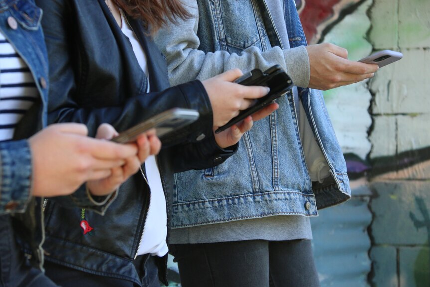 Three teenagers, all on their phones, lean against a wall with graffiti on it.