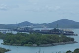 The coast with a shipping boat. There are trees in the picture before the ship and a small mountain in the background.