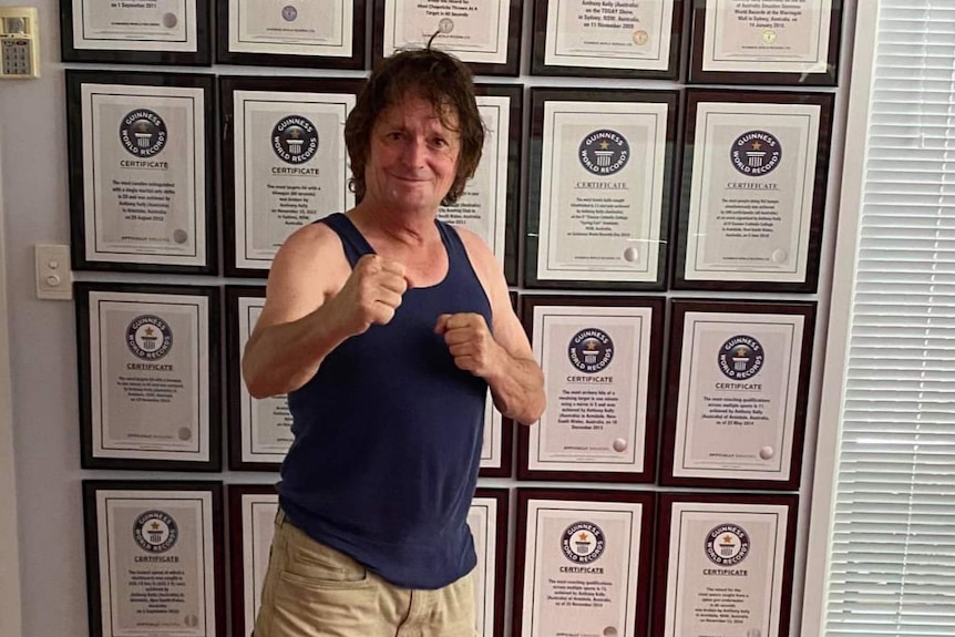 A man in a blue singlet and beige shorts stands in front of a wall of certificates, posing with his fists