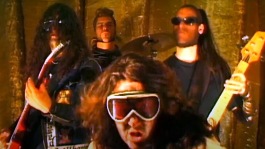 A blurry image of four men with long hair wearing goggles and glasses and playing instruments in a brown room