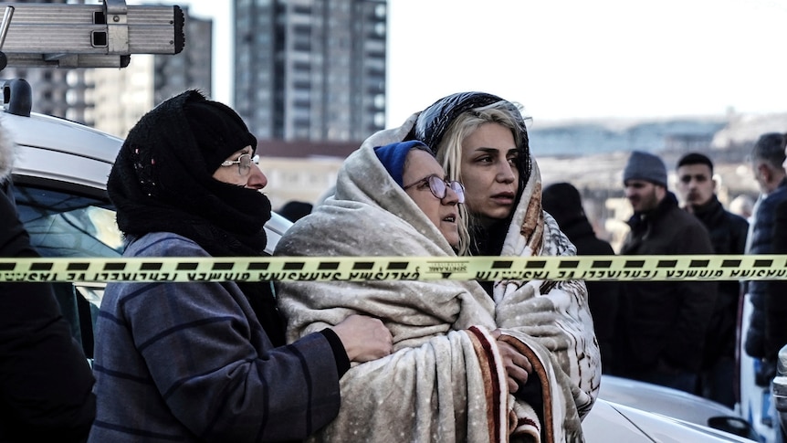 Three women huddle together in blankets, standing behind emergency tape. 