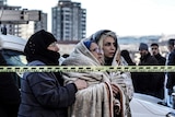 Three women huddle together in blankets, standing behind emergency tape. 