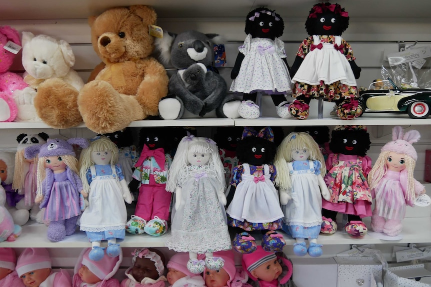 Golliwogs, rag dolls and teddy bears sitting on a shelf inside a shop.