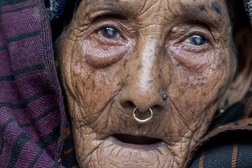 A close up portrait of an elderly Nepalese woman who has a stud and a hoop earring in her nose and wears a head wrap