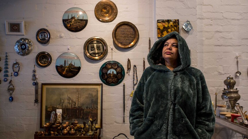 Onur wears a green fake fur hooded coat and stands in front Turkish wall decorations