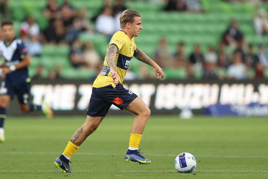 A soccer player wearing yellow and navy blue dribbles the ball along the grass during a game