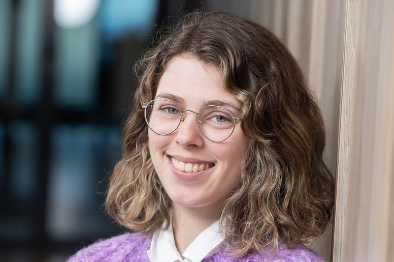 A young girl wearing a purple jumper and glasses smiles at the camera.