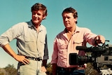 An old coloured photo of two smiling men in the outback, blue sky, red earth, stand next to ta film camera. Both w