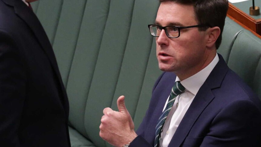 A man with glasses, sitting alongside a woman on green parliamentary benches, gives a thumbs up as another man walks past.