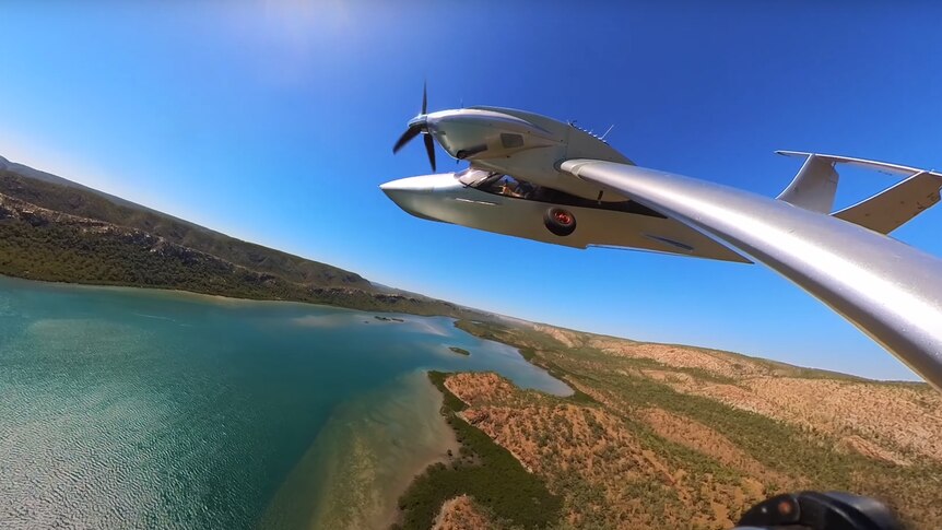 A silver seaplane flies over water and arid landscape, in front of blue sky.