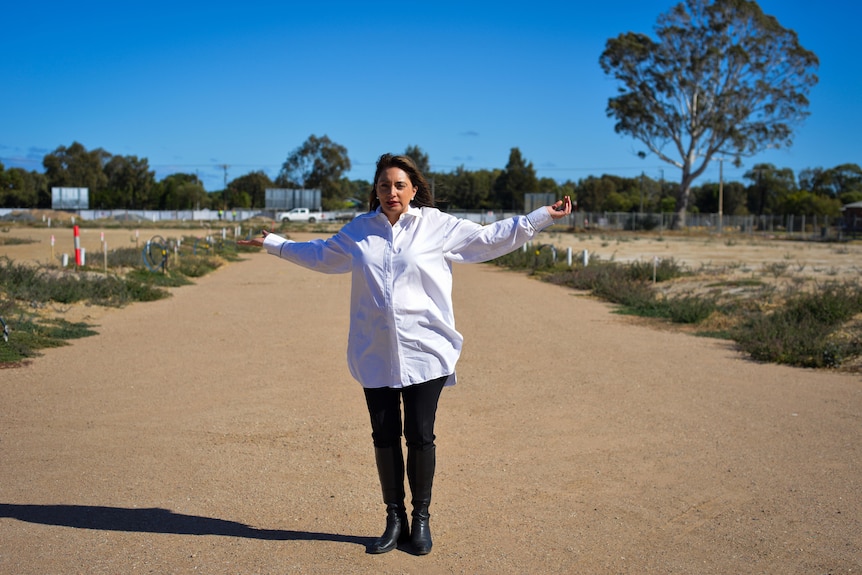 A woman wearing a white shirt and black pants stands on a vacant block with her arms outstretched