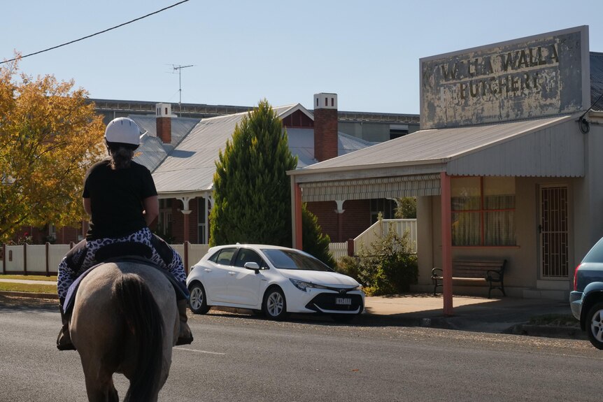 A woman rides a horse on a quiet street with an old butchery in the background. 