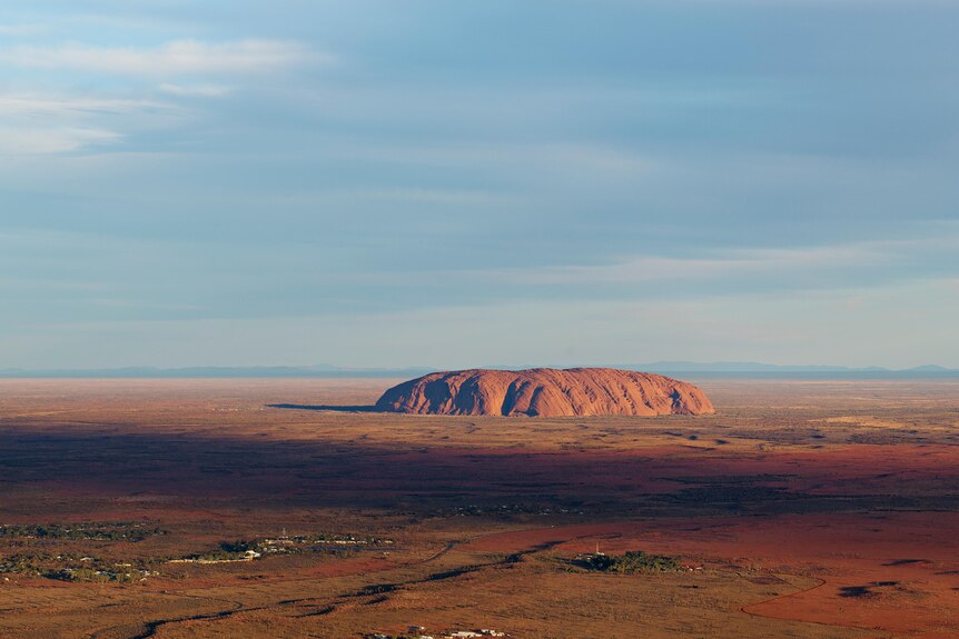 A wide aerial view of Uluru and its surrounding landscape on a sunny day.