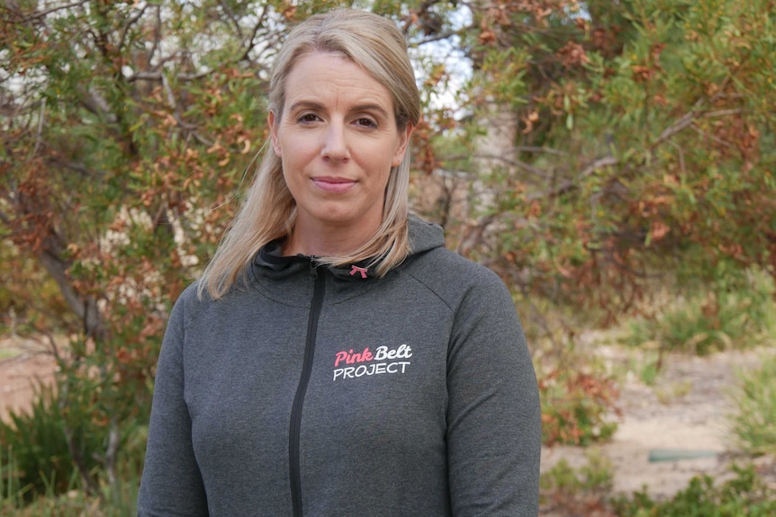 A woman in a grey jacket stands in front of a tree.