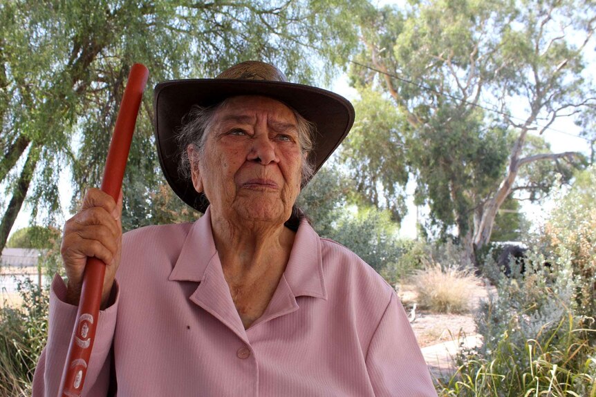 An older Aboriginal woman holds a staff and looks past the camera.