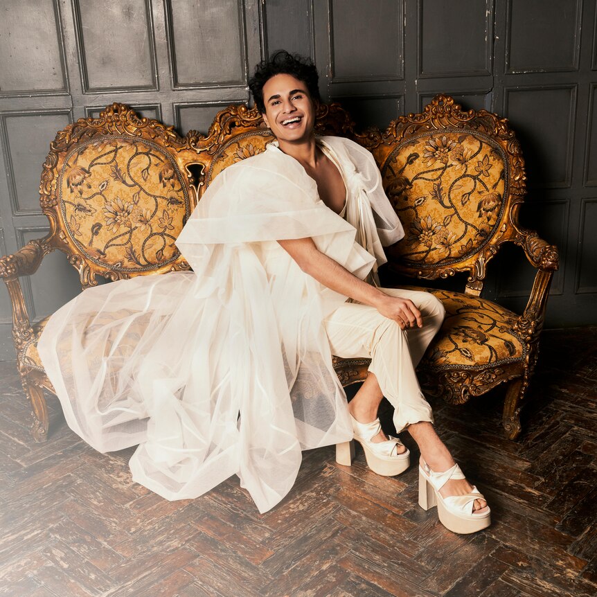 Male soprano Samuel Mariño reclining on a lounge and smiling in a white gown.